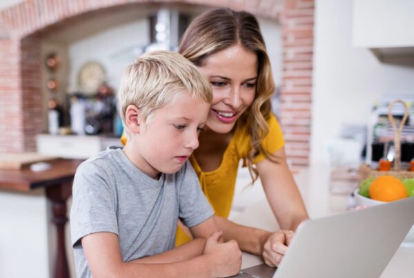 A young woman and a boy are sitting together, looking at a laptop. They are indoors in a kitchen setting, with light-colored walls around them. The woman is dressed casually, and both have focused expressions as they engage with the screen.