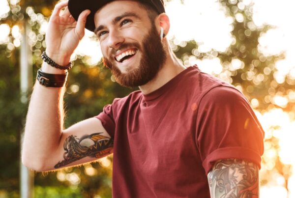 Man in red t-shirt, baseball hat wearing ear pods smiling with sleeve tattoos on both arms