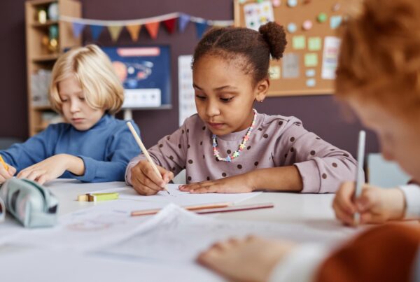Alt text: A group of children sitting at a table in a classroom, focused on writing on paper. The students, including both girls and boys, are engaged in learning activities.