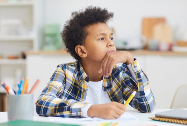 A child sitting at a desk in a classroom, surrounded by office supplies and furniture, focused on learning. The setting includes a wall in the background.