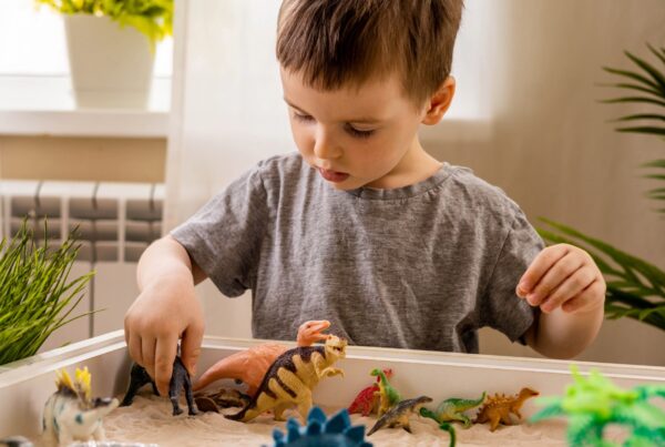 A toddler playing with colorful toys indoors, wearing casual clothing. The scene includes a birthday cake in the background, a wall with decorations, and a small plant nearby. The child has a joyful expression on their face as they engage with the toys.