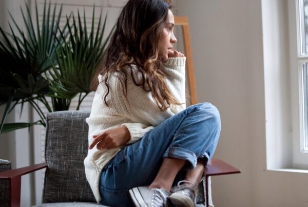 A woman sitting on a chair indoors, wearing jeans. The room has a wall with a window, furniture including a couch and a vase, and a houseplant nearby.