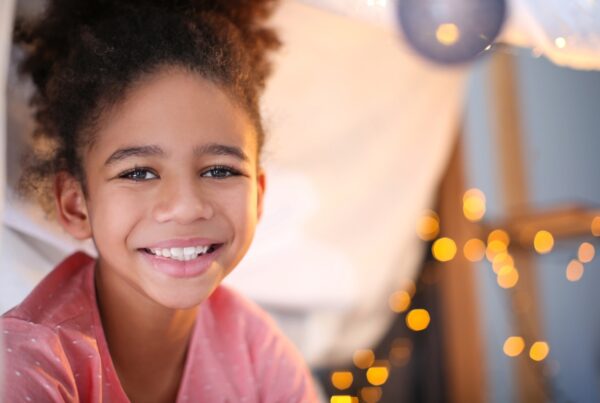 Close-up portrait of a young woman with a joyful smile, showing her teeth. She has well-groomed eyebrows and is indoors wearing casual clothing.