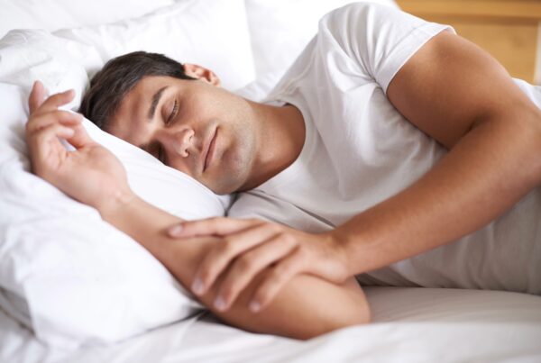 A person sleeping peacefully on a bed, covered with a blanket, in a cozy indoor setting. The individual is wearing comfortable clothing and appears relaxed.